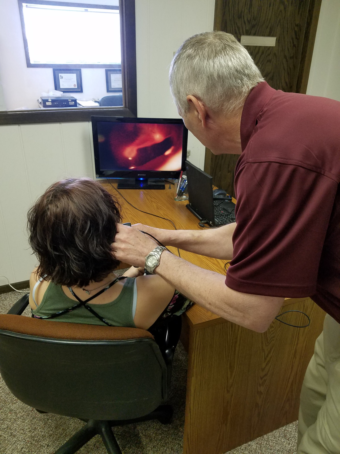 behind hearing specialist and patient at desk while he uses an instrument to examine patient ear