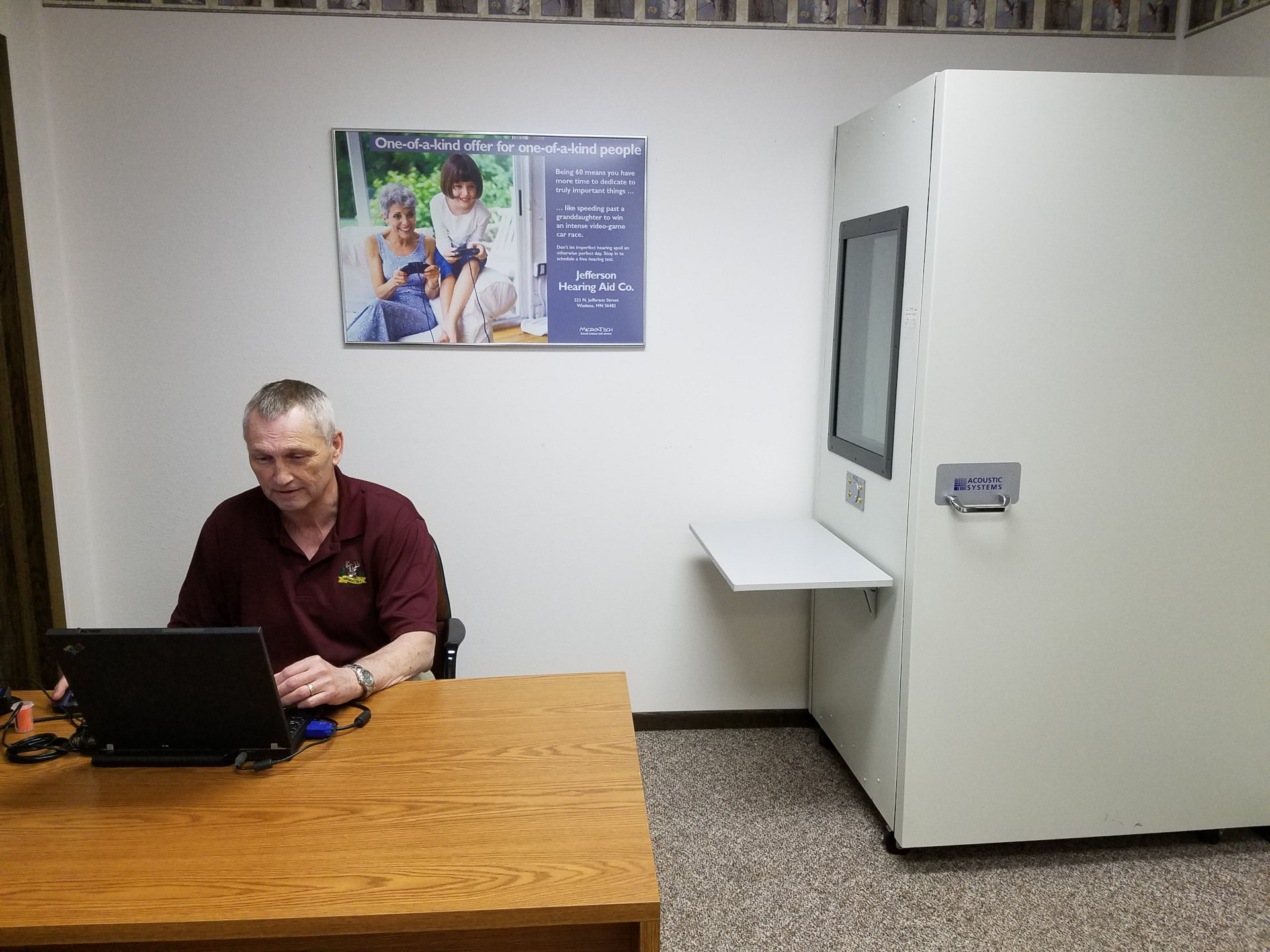 hearing specialist working at laptop with hearing test booth visible in background